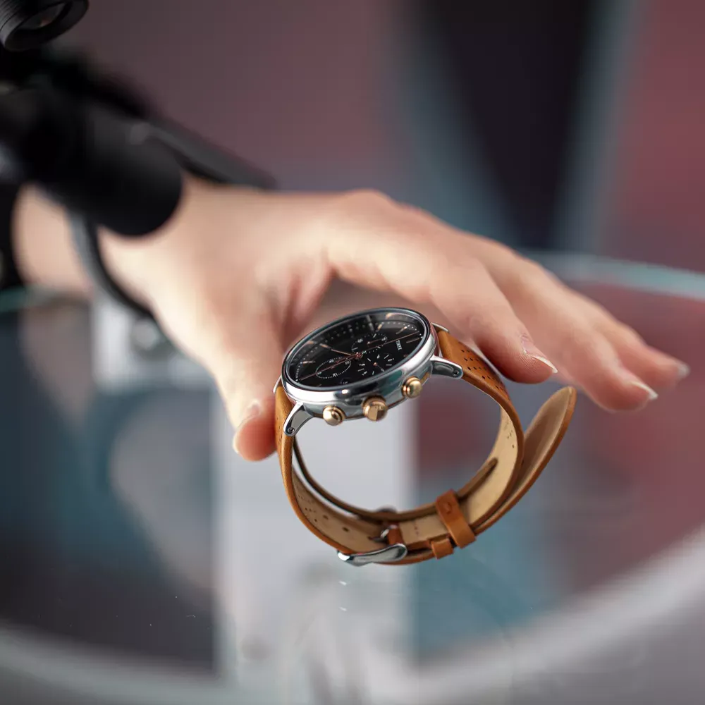 Mano posicionando reloj de hombre con correa de cuero marrón sobre superficie de vidrio